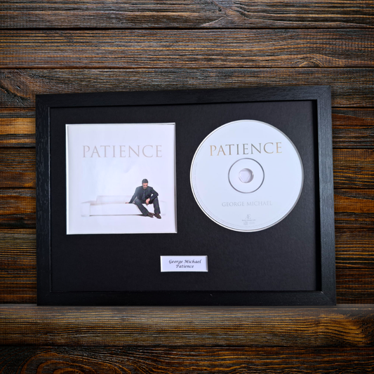 A framed music memorabilia display featuring George Michael's album 'Patience' on a rustic dark wood background. The black frame contains the square album cover, a silver CD, and a small white nameplate reading 'George Michael - Patience'.