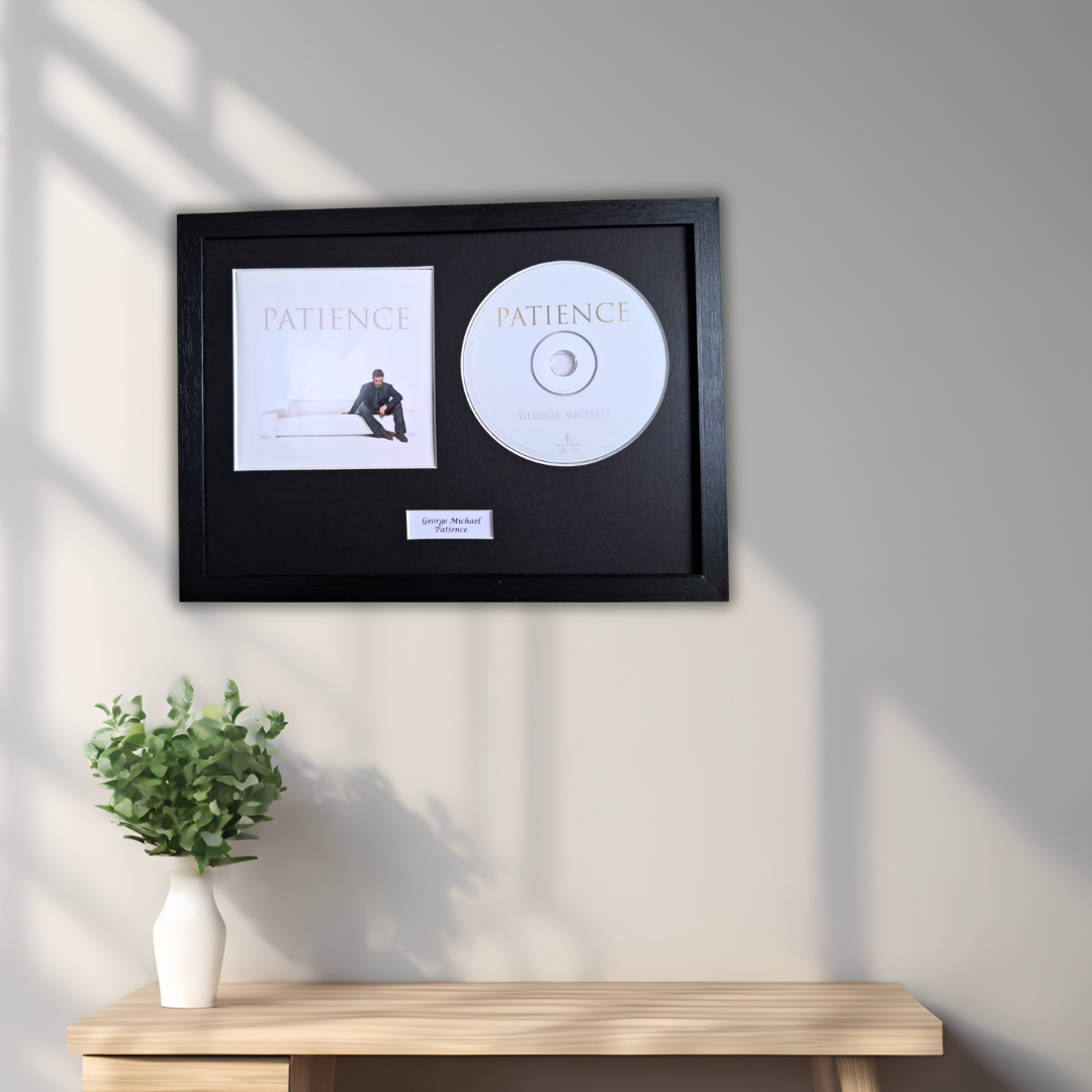 A framed music display featuring a black frame with a Patience album cover print and a white CD, mounted on a light gray wall above a wooden console table with a small green plant in a white vase.