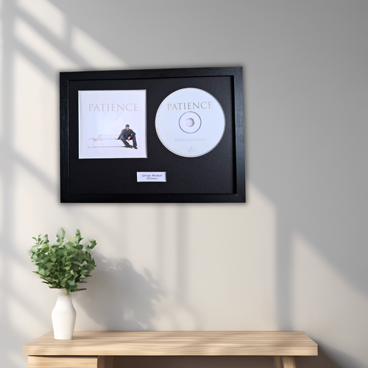 A framed music display featuring a black frame with a Patience album cover print and a white CD, mounted on a light gray wall above a wooden console table with a small green plant in a white vase.