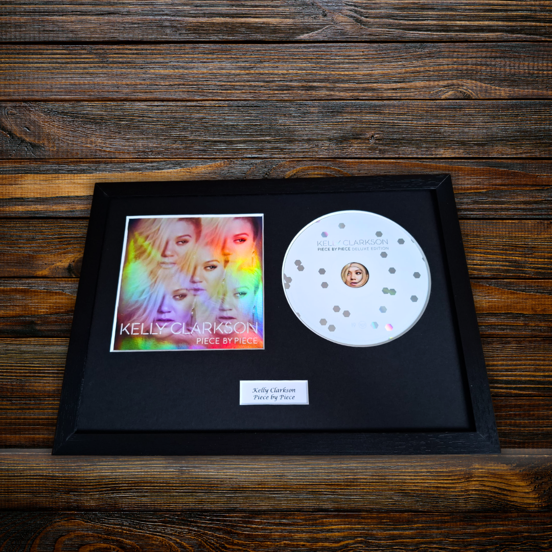 A framed Kelly Clarkson 'Piece by Piece' CD display on a dark wood background. The black frame contains the CD, a printed album cover insert, and a small white label.