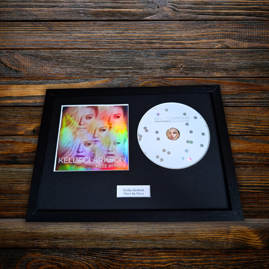 A framed Kelly Clarkson 'Piece by Piece' CD display on a dark wood background. The black frame contains the CD, a printed album cover insert, and a small white label.