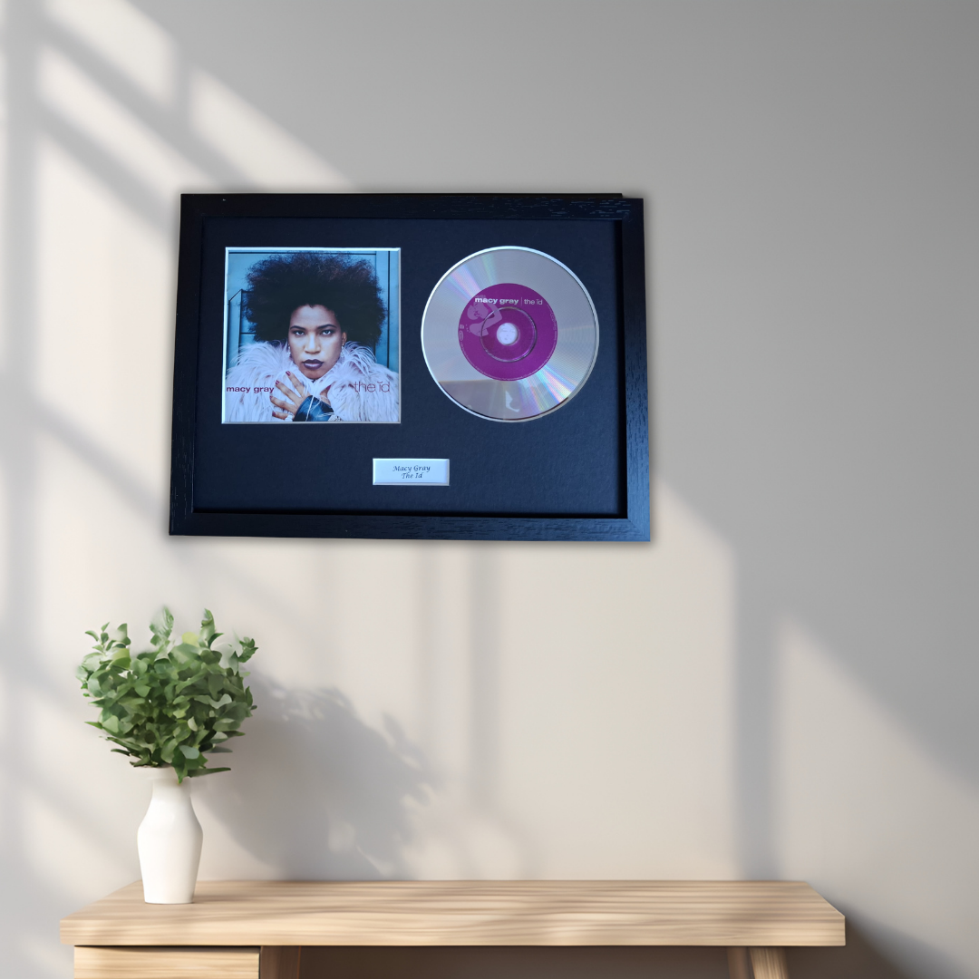 A framed music memorabilia display hanging on a light gray wall above a wooden console table. The black frame contains a small portrait photo, a purple-and-silver CD, and a small plaque.