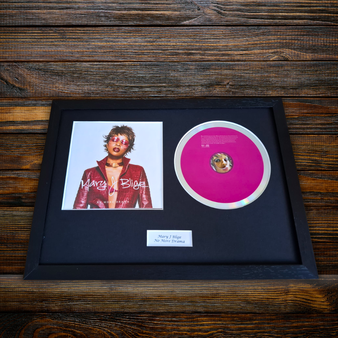 A framed Mary J Blige - No More Drama sign on a dark wood background, featuring a black frame with a black mat, a portrait photo of Mary J Blige, a bright pink record disc, and a small white label.