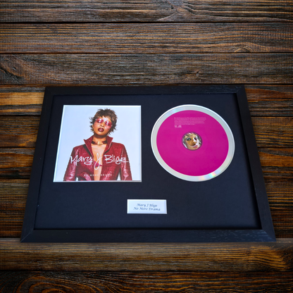 A framed Mary J Blige - No More Drama sign on a dark wood background, featuring a black frame with a black mat, a portrait photo of Mary J Blige, a bright pink record disc, and a small white label.