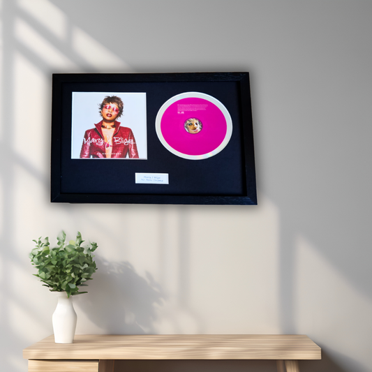 A framed music memorabilia piece displayed on a light gray wall above a wooden console table with a small potted plant. The black frame contains a portrait photo of a man in a red jacket and a bright pink record disc with a white border.