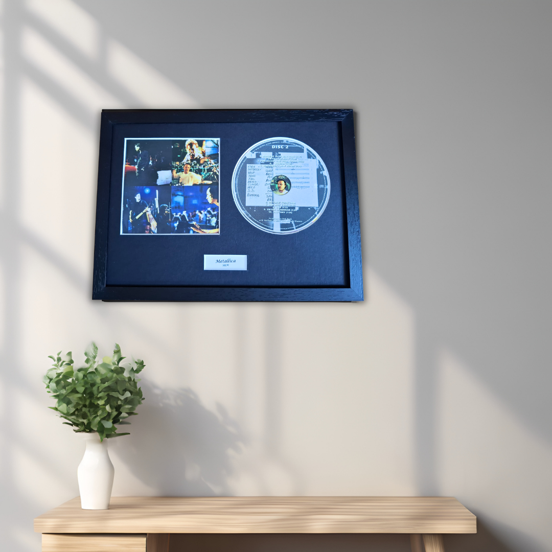 A framed black record plaque featuring a small photo collage and a circular silver-toned disc, mounted on a light gray wall above a wooden console table with a white vase of green leaves.