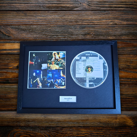A framed metal record display featuring a circular transparent CD, a four-photo collage, and a small 'Metallica' plaque on a black mat, set against a dark wood background.