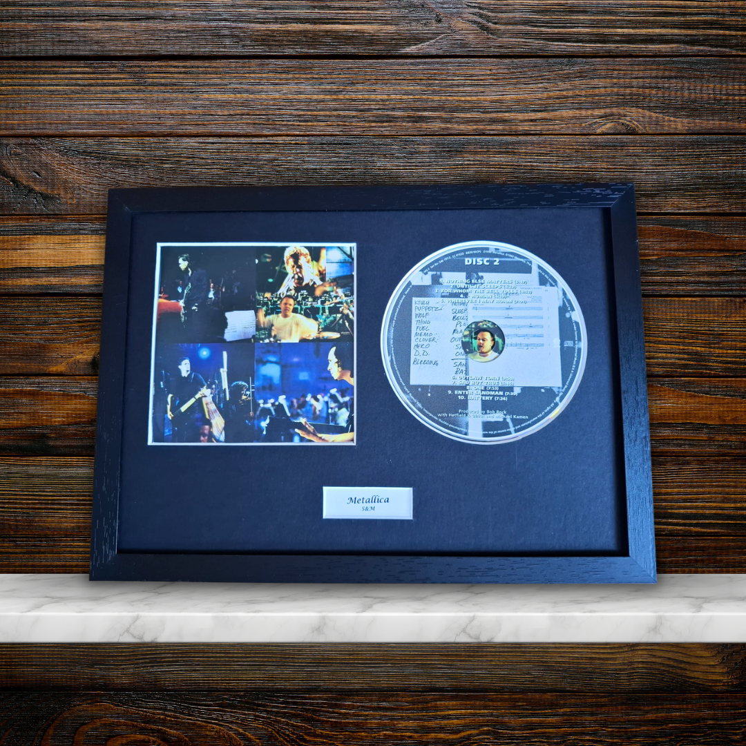 A framed Metallica 'S&M' display featuring a clear acrylic disc, a photo collage, and a nameplate inside a black frame, set against a dark wood background.