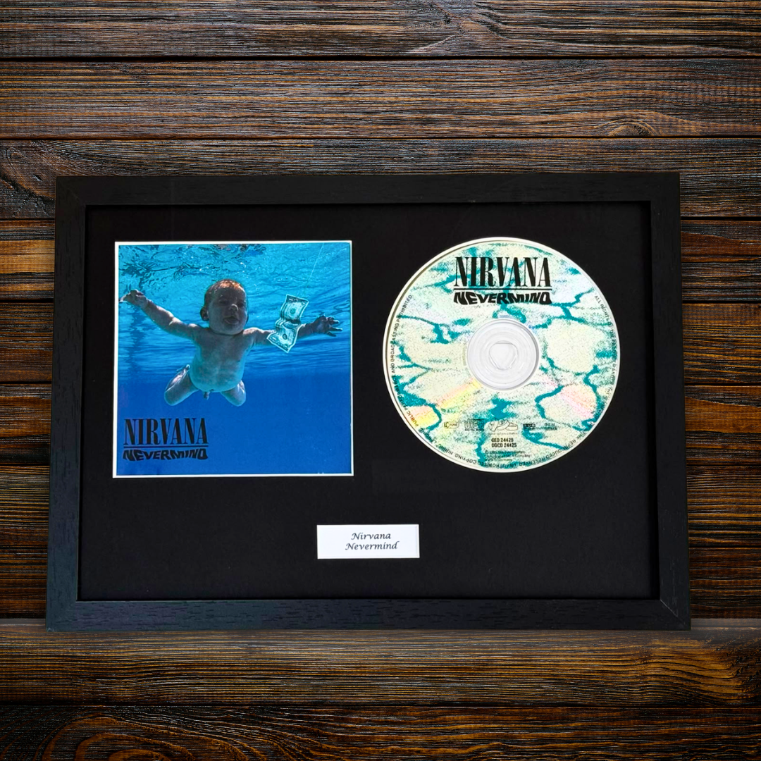 A framed Nirvana 'Nevermind' album display on a dark wood background, featuring the album cover photo, a CD disc, and a small label in a black frame.