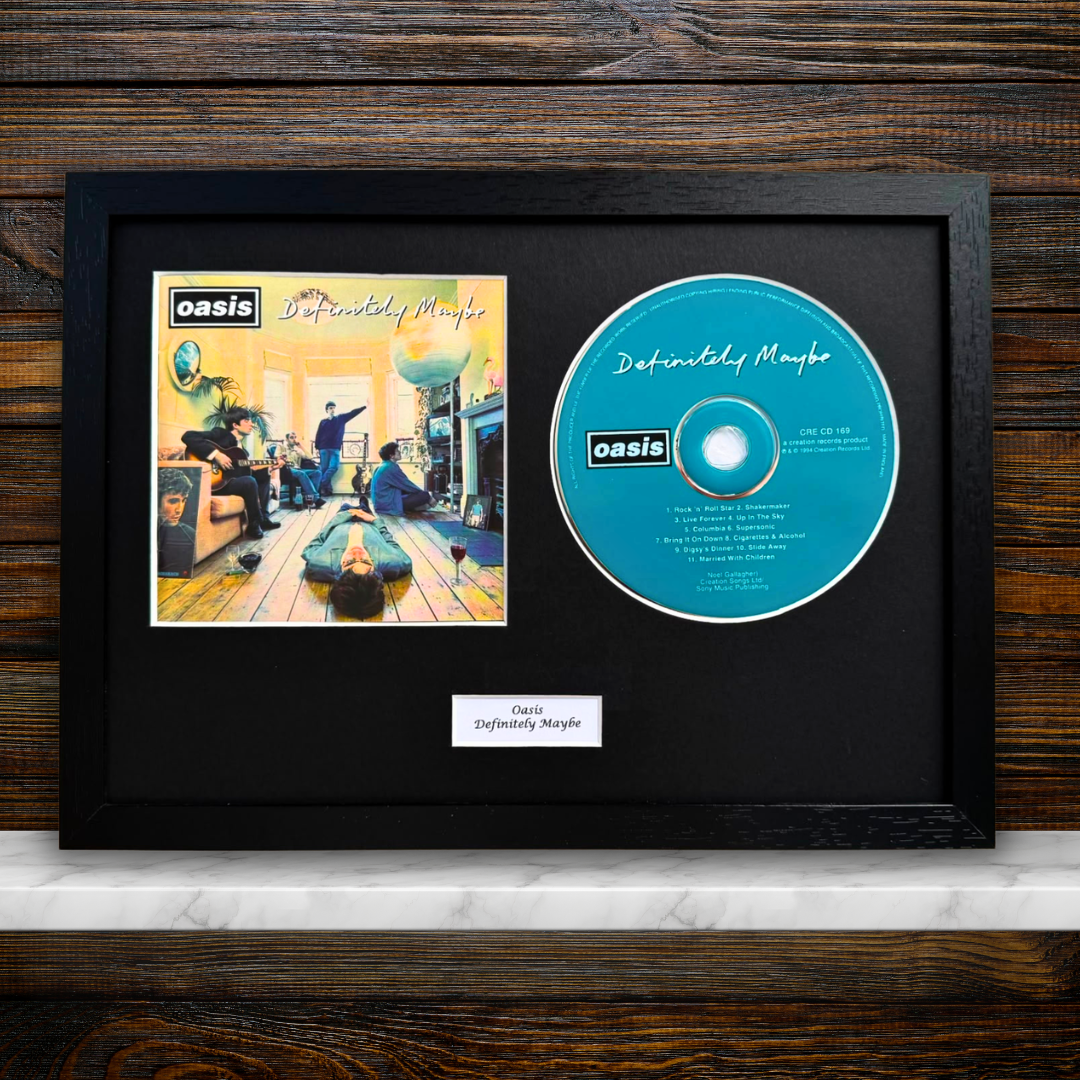 A framed Oasis 'Definitely Maybe' album display featuring the printed album cover and a blue CD on a black mat, with a small label below, set against a dark wood background.