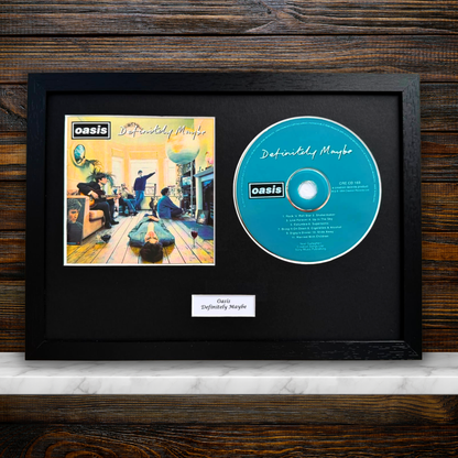 A framed Oasis 'Definitely Maybe' album display featuring the printed album cover and a blue CD on a black mat, with a small label below, set against a dark wood background.