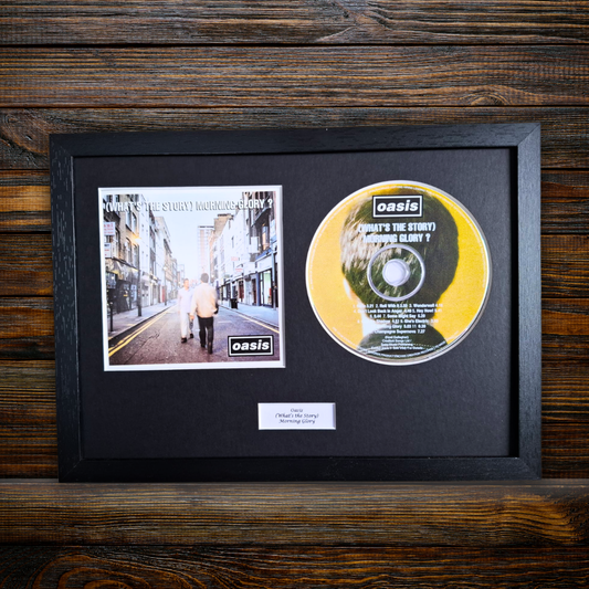 A framed Oasis '(Whats The Story) Morning Glory?', framed CD Artwork including the album cover and the yellow/green CD, mounted on a black mat inside a black frame against a dark wood background.