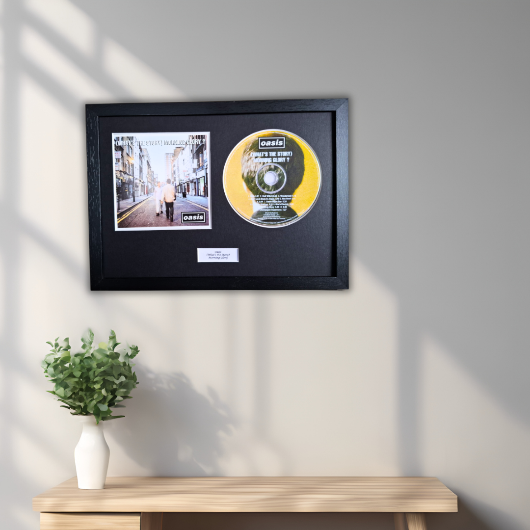 A framed CD single display mounted on a light gray wall above a wooden console table with a small white vase of green leaves. The black frame contains a printed street scene photo and a yellow/gold CDof Oasis (What's The Story) Morning Glory?