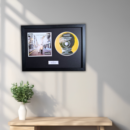 A framed CD single display mounted on a light gray wall above a wooden console table with a small white vase of green leaves. The black frame contains a printed street scene photo and a yellow/gold CDof Oasis (What's The Story) Morning Glory?