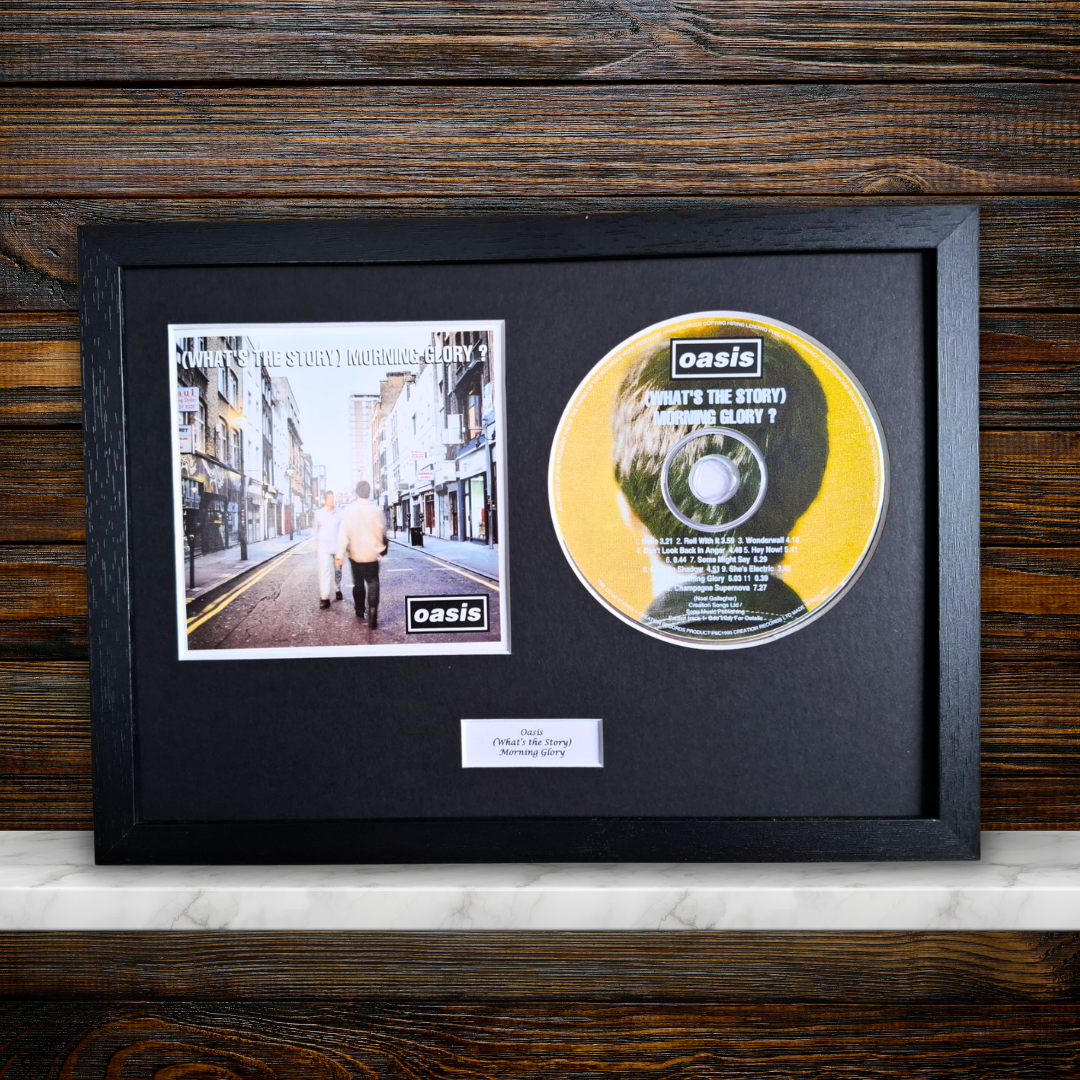 A framed Oasis '(What's The Story) Morning Glory?' CD album, featuring the black album cover photo and the yellow CD disc inside a black shadowbox frame, displayed on a white marble shelf against a dark wood background.