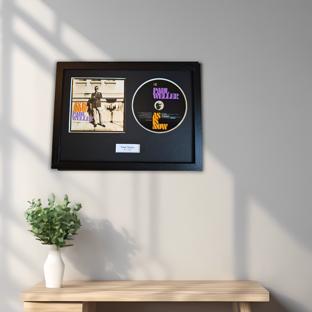 A framed music memorabilia display hanging on a light gray wall above a wooden console table. The black frame contains a small album cover print, a circular black disc with purple and orange text, and a small white plaque.