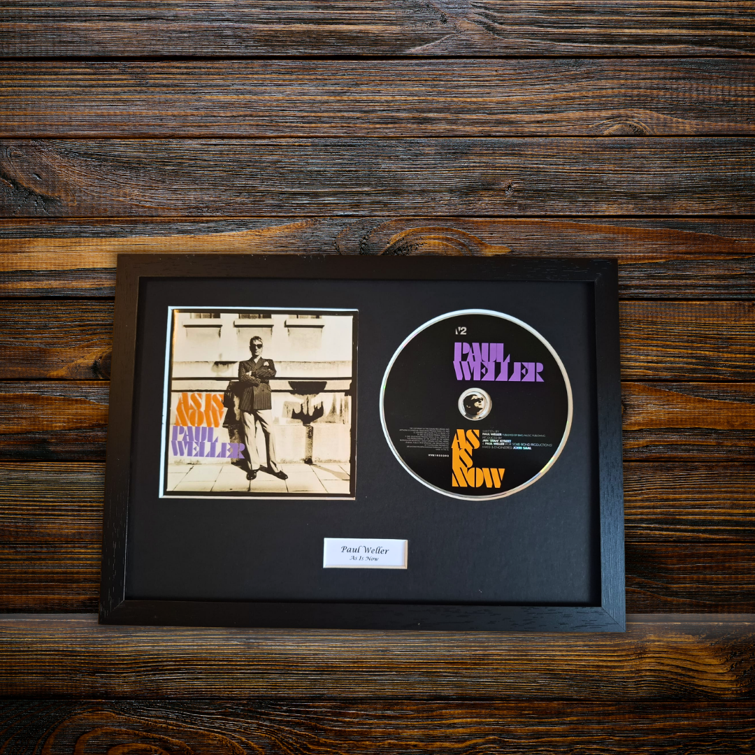 A framed music memorabilia piece featuring a vintage CD single and a printed album cover mounted on a black mat inside a black frame, displayed against a dark wood plank background.