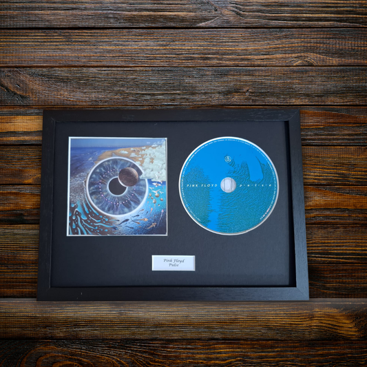 A framed music memorabilia piece featuring a blue CD and a square printed album art print inside a black frame, mounted on a dark wood background. A small label at the bottom reads 'Pink Floyd - Pulse'