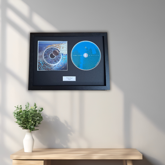 A framed music award display featuring a blue vinyl record and a square photo print mounted on a black background, hung on a light gray wall above a wooden console table with a small white vase of greenery.