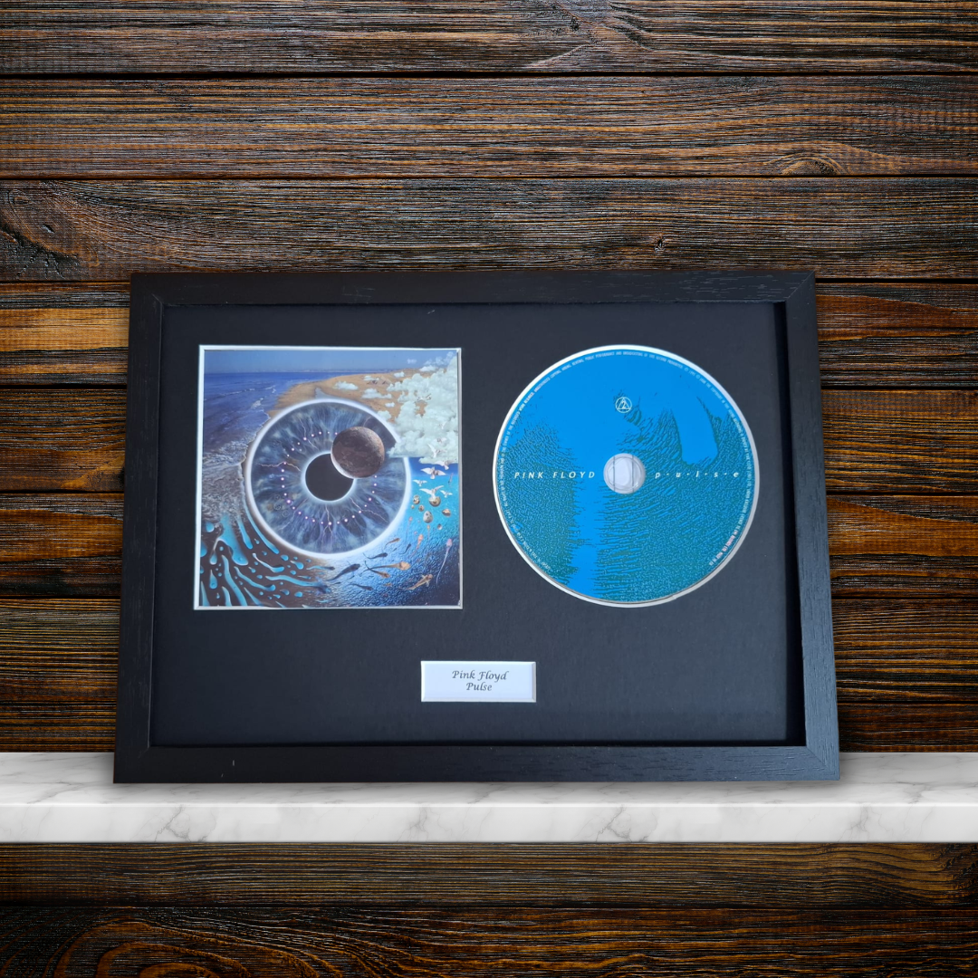 A framed Pink Floyd 'Pulse' collectible display featuring a blue original CD, a printed album cover, and a small label on a black mat, set against a dark wood background.