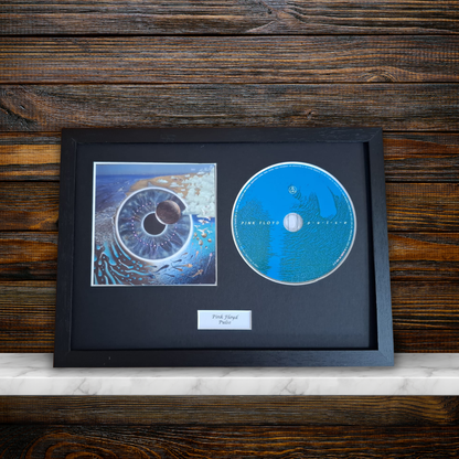 A framed Pink Floyd 'Pulse' collectible display featuring a blue original CD, a printed album cover, and a small label on a black mat, set against a dark wood background.