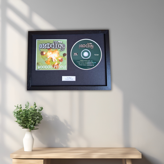 A framed music memorabilia display mounted on a light gray wall above a wooden console table. The black frame contains a square album cover insert and a green CD disc with the title 'Voodoo People' by 'the Prodigy'.