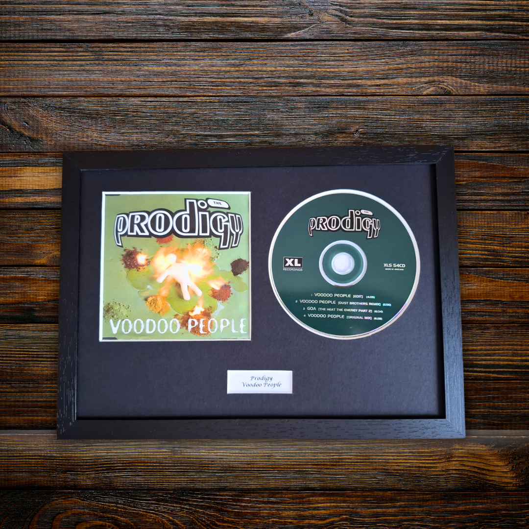 A framed music display featuring the Prodigy 'Voodoo People' CD and album cover inside a black shadowbox frame, placed on a dark wood background.