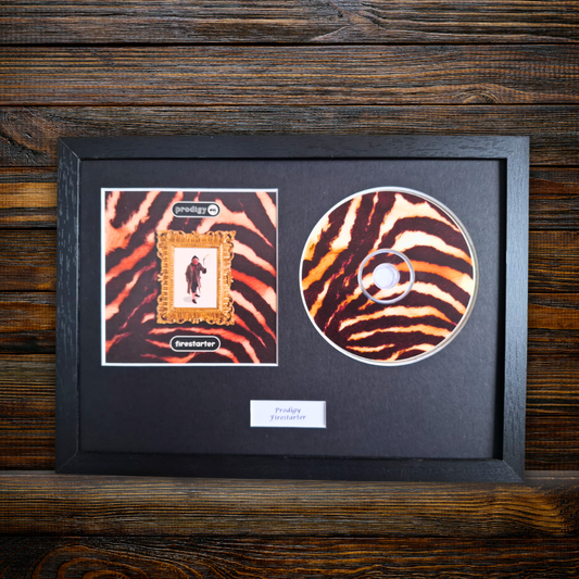 A framed music memorabilia display featuring a CD and a printed album cover insert on a black mat, set against a dark wood plank background. The album cover has a zebra-stripe pattern with a central gold-bordered portrait frame, and the CD also has a matching zebra pattern.