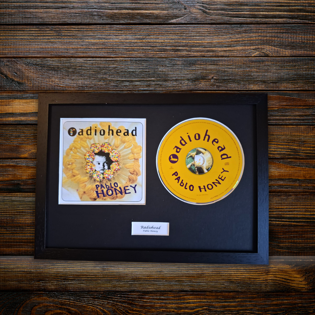 A framed Radiohead 'Pablo Honey' album display on a dark wood background, featuring the square album cover and a yellow CD inside a black frame with a small label below.