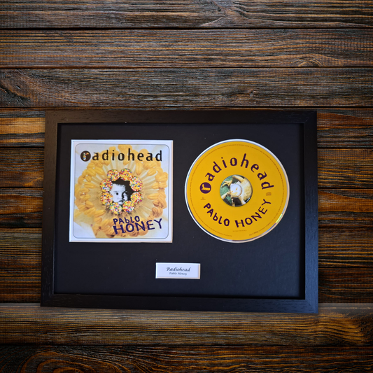 A framed Radiohead 'Pablo Honey' album display on a dark wood background, featuring the square album cover and a yellow CD inside a black frame with a small label below.