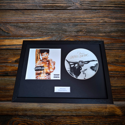 A framed memorabilia display on a dark wood background, featuring a black album cover print, a white signed CD, and a small white plaque 