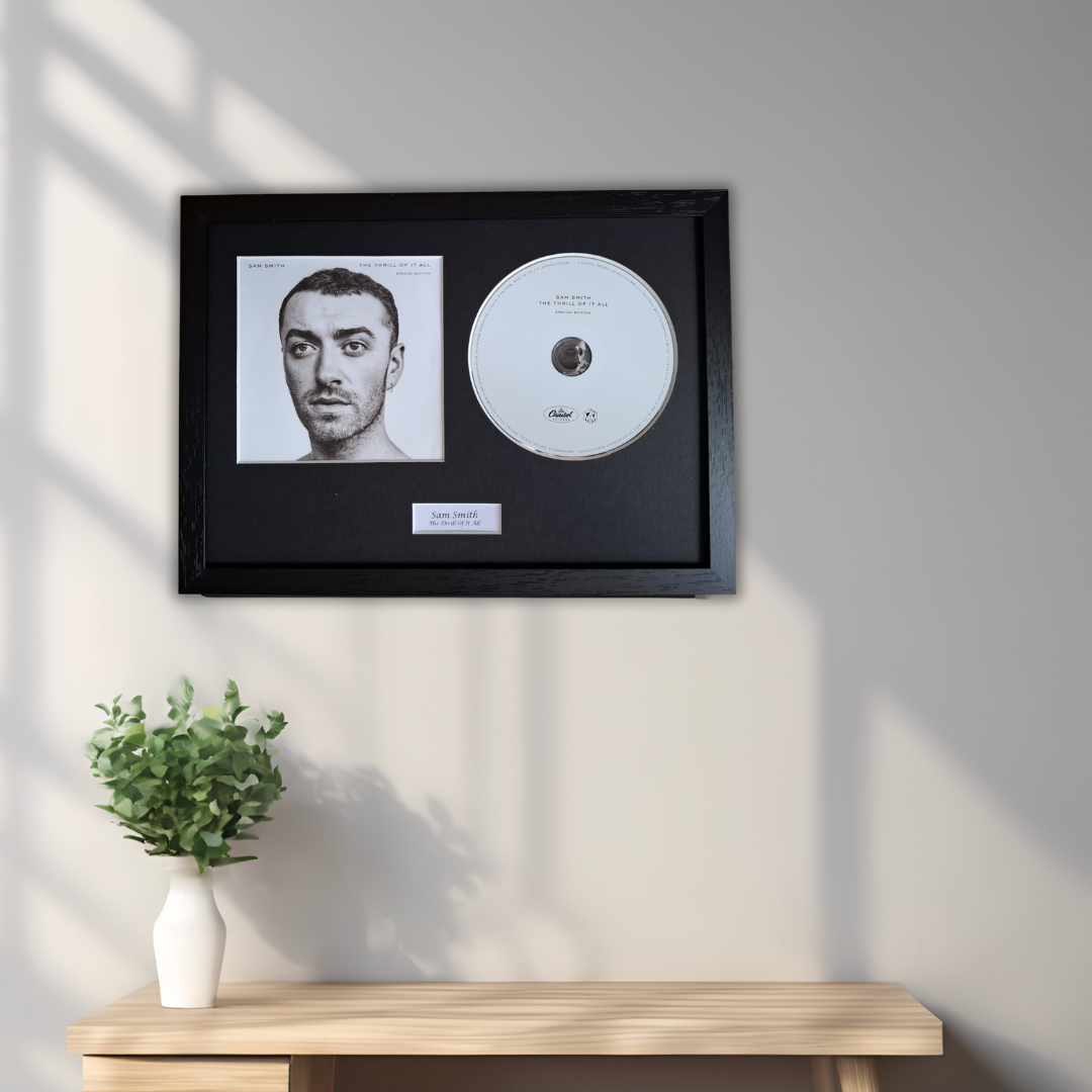A framed black-and-white portrait of Sam Smith with a white CD and small plaque inside a black frame, mounted on a light gray wall above a wooden console table with a white vase of green leaves.
