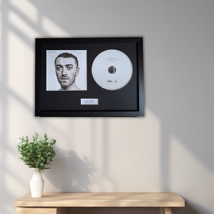A framed black-and-white portrait of Sam Smith with a white CD and small plaque inside a black frame, mounted on a light gray wall above a wooden console table with a white vase of green leaves.