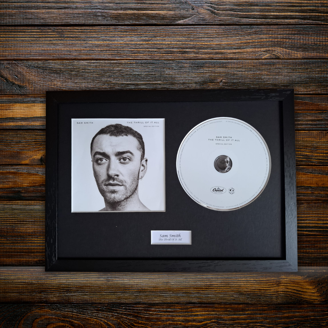 A framed music memorabilia piece featuring a black picture-frame display with a black mat, a printed portrait of Sam Smith, a silver CD, and a small nameplate reading 'Sam Smith - The Thrill of It All' on a rustic dark wood background.