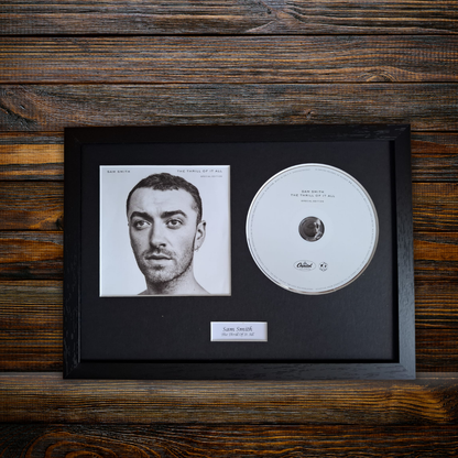 A framed music memorabilia piece featuring a black picture-frame display with a black mat, a printed portrait of Sam Smith, a silver CD, and a small nameplate reading 'Sam Smith - The Thrill of It All' on a rustic dark wood background.