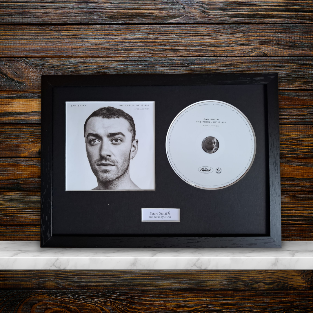 A framed music memorabilia piece featuring a black-and-white portrait print of Sam Smith and a silver-colored CD inside a black frame, mounted on a black mat against a dark wood plank wall with a white marble shelf visible below.