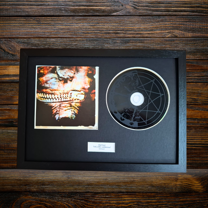 A framed music memorabilia display featuring a CD with a pentagram design and a small printed album insert, mounted in a black frame against a dark wood background.