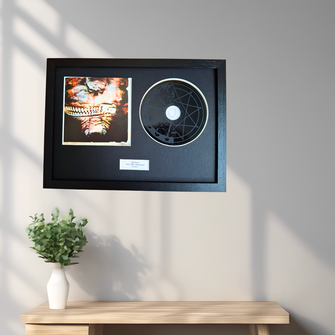 A framed black record plaque mounted on a light gray wall above a wooden console table with a small white vase of greenery. The frame contains a square printed artwork on the left and a circular black vinyl record on the right, with a small white label below.