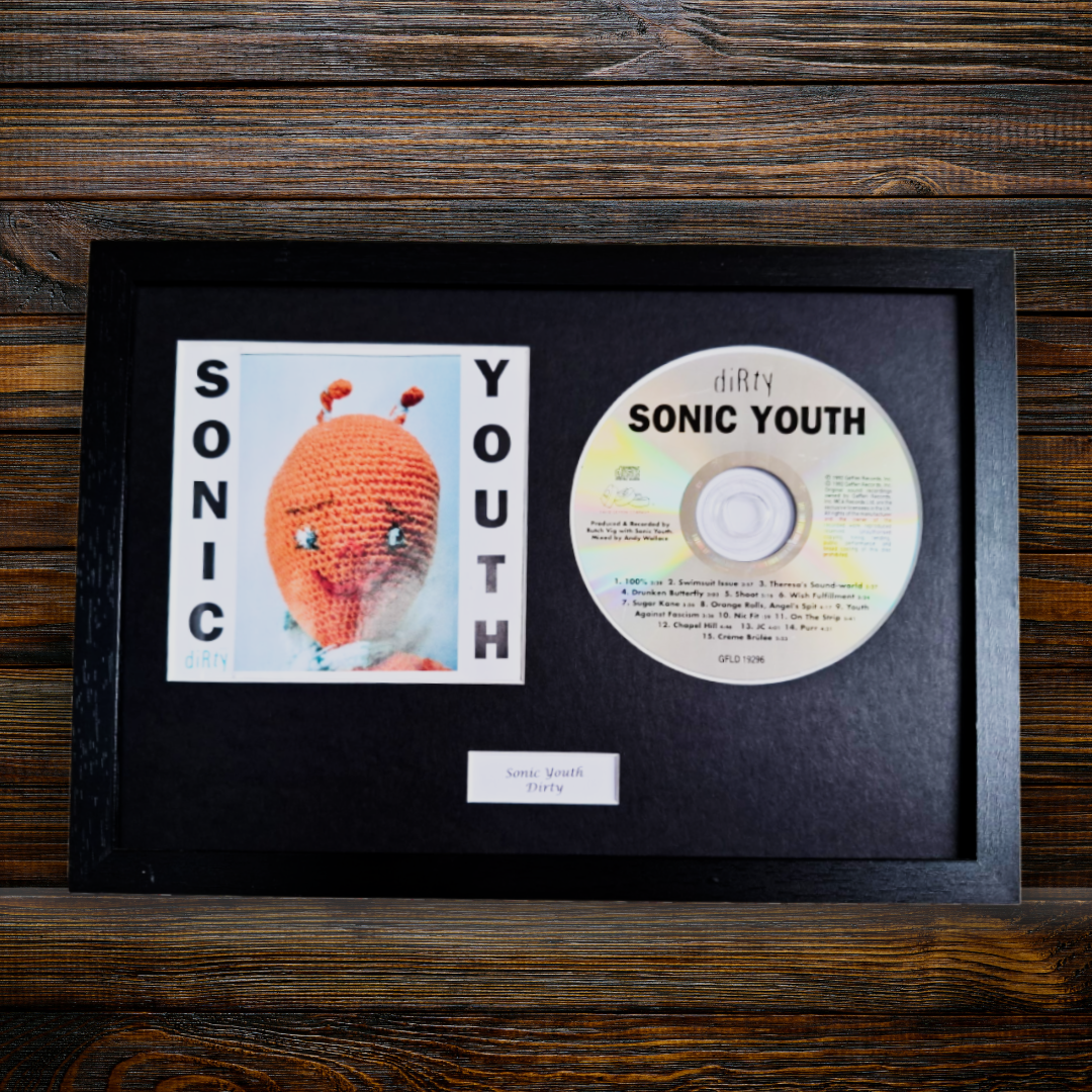 A framed Sonic Youth - Dirty photo display featuring a black frame with a black mat, a printed album photo of the crocheted character, the CD, and a small label on a dark wood background.