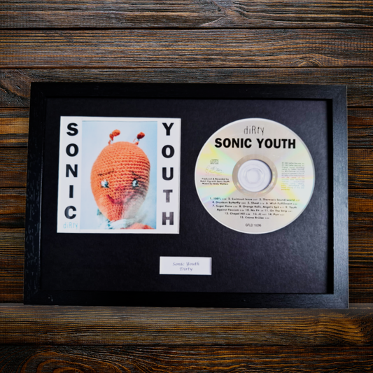 A framed Sonic Youth - Dirty photo display featuring a black frame with a black mat, a printed album photo of the crocheted character, the CD, and a small label on a dark wood background.