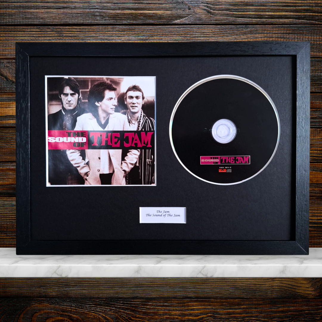 A framed music memorabilia display featuring a black CD with a The Jam label sticker, alongside a printed band photo and a small plaque, all mounted on black mat inside a black frame against a wood panel background.