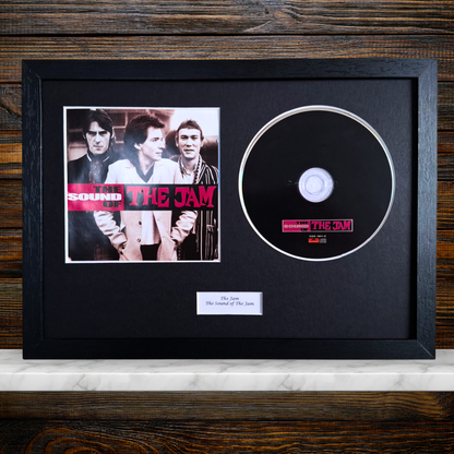 A framed music memorabilia display featuring a black CD with a The Jam label sticker, alongside a printed band photo and a small plaque, all mounted on black mat inside a black frame against a wood panel background.