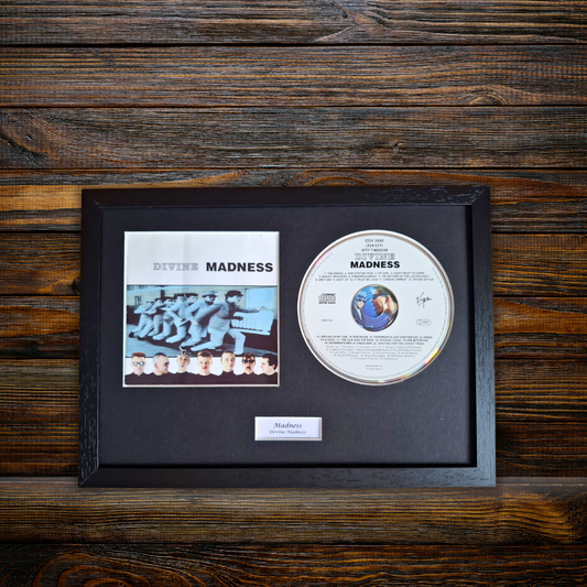 A framed and signed copy of the band Divine Madness 'Madness' album, featuring the black album cover insert and a signed CD inside a black wooden frame, displayed on a dark wood background.