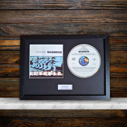 A framed Divine Madness CD album display featuring the original CD, printed cover art, and a small plaque on a black mat inside a dark wood frame, set against a dark wood panel background.