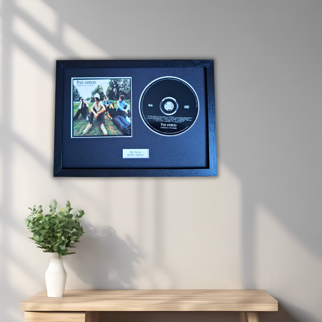A framed music memorabilia display hanging on a light gray wall above a wooden console table. The frame contains a small square album cover photo and a black CD mounted on a dark navy backing, with a small white plaque below.